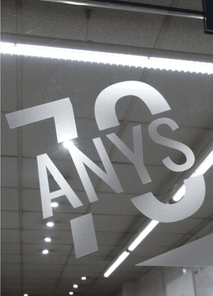 Vinilo plateado con el texto "70 anys" en letras grandes y tipografía moderna pegado en el escaparate interior del taller Contell Marcs de Barcelona, celebrando los 70 años de experiencia en enmarcación artesanal.