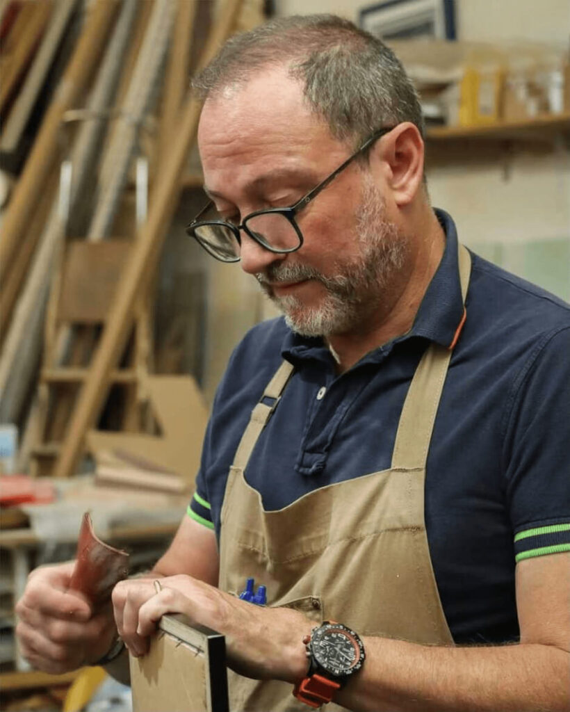 Enmarcador artesano con delantal aplicando acabado a mano en una moldura de marco negro en el taller Contell Marcs de Barcelona, rodeado de perfiles de madera y materiales de enmarcación.
