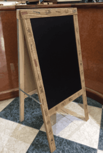 Caballete tipo A con pizarra negra y marco de madera artesanal fabricado por Contell Marcs Barcelona, con estampado estilo caja de transporte frágil en inglés y español, sobre suelo de mármol verde de local comercial.