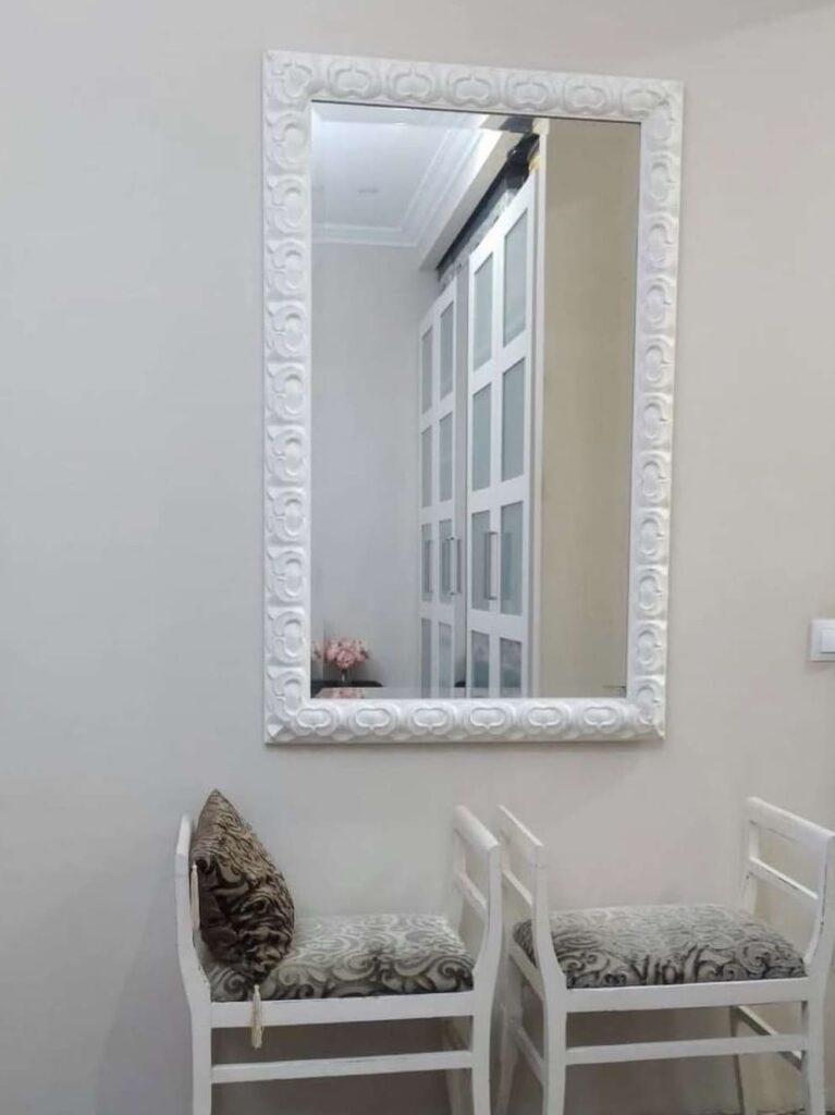 Espejo rectangular con marco blanco de relieve decorativo colgado en pared de habitación, con dos sillas tapizadas debajo, fabricado por Contell Marcs.