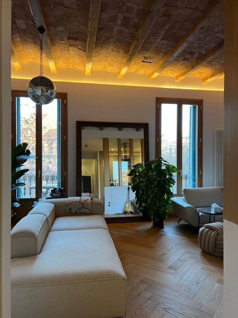 Gran espejo con marco de madera oscura tallada, apoyado en la pared de un salón moderno con techo bóveda catalana, sofá beige y plantas, enmarcado por Contell Marcs.