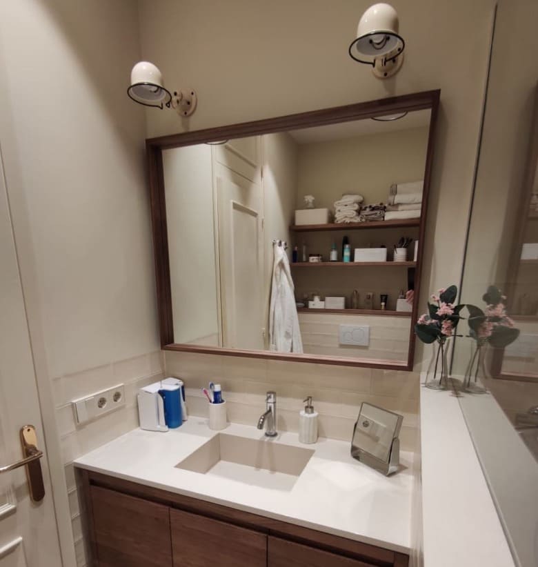 Espejo rectangular con marco de madera oscuro a medida instalado sobre lavabo empotrado en baño moderno con apliques de pared blancos y estantes con productos, fabricado por Contell Marcs.
