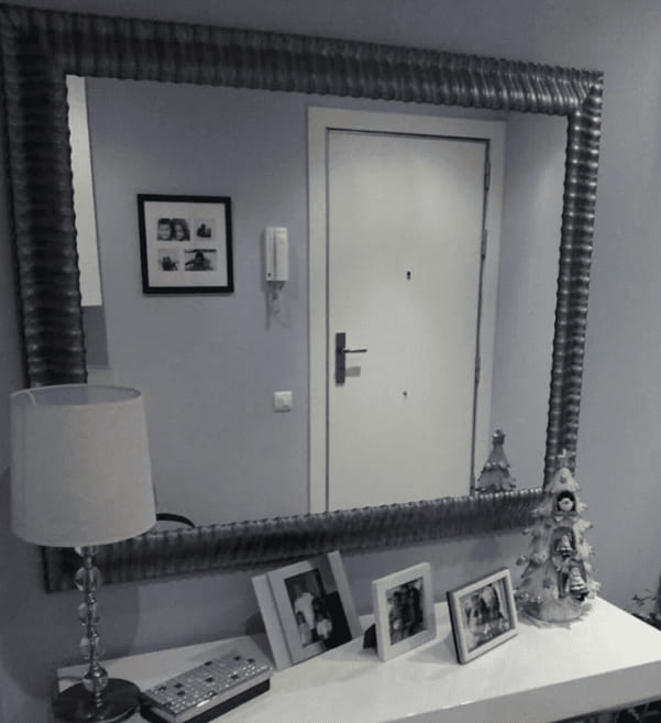 Espejo de gran formato con marco de madera plateado de moldura ondulada y textura metálica instalado en recibidor de hogar, sobre consola blanca con portafotos y lámpara, fabricado a medida por Contell Marcs.