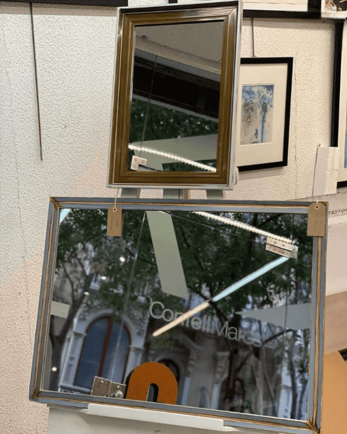 Dos espejos a medida expuestos en el escaparate del taller Contell Marcs de Barcelona: uno con marco plateado efecto espejo y otro con marco vinatge, reflejando la fachada de la tienda.