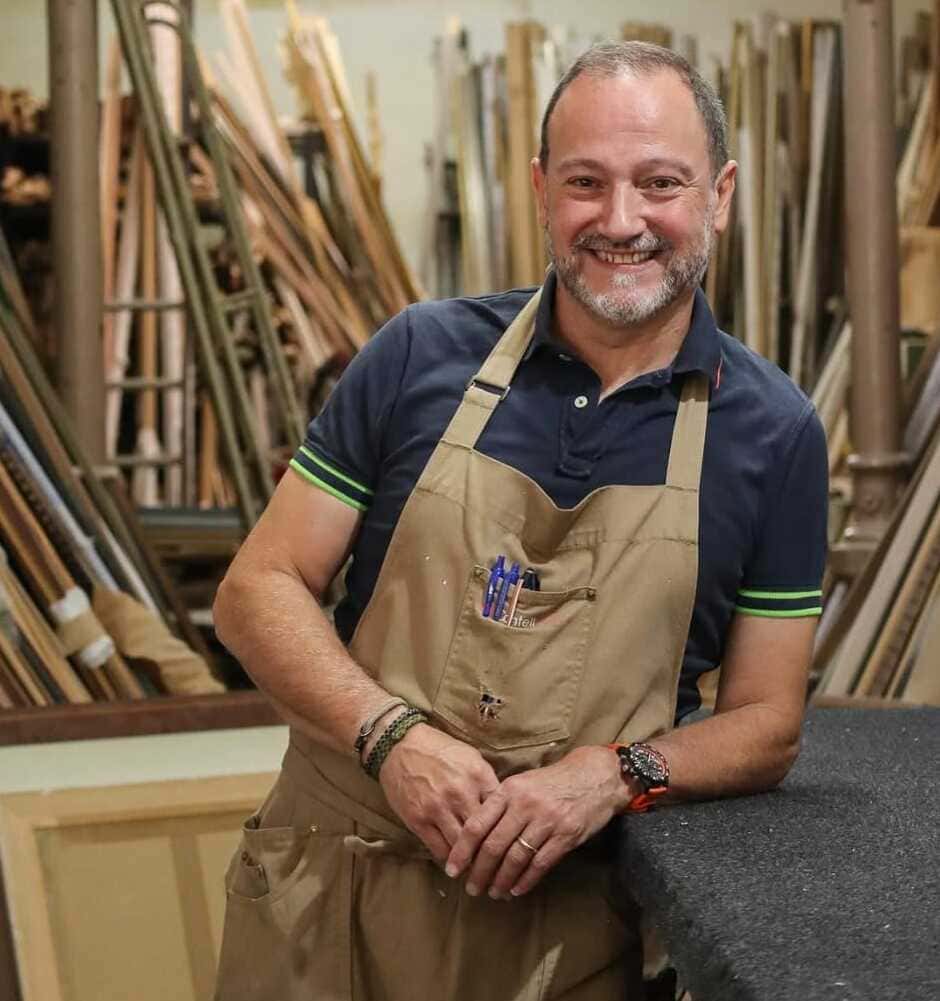 Retrato del maestro enmarcador de Contell Marcs sonriendo con delantal en su taller artesanal de Barcelona, rodeado de cientos de molduras y perfiles de madera de diferentes acabados y estilos.