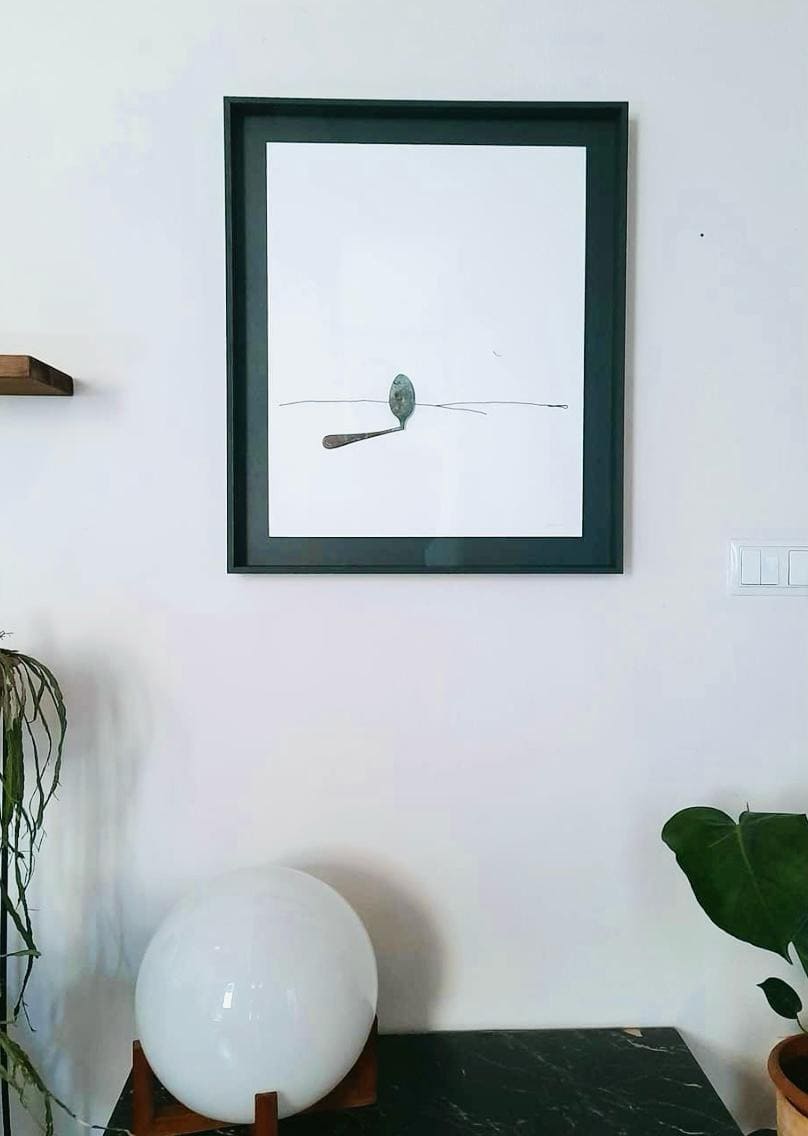 Dibujo minimalista en tinta negra sobre fondo blanco con una figura abstracta y línea horizontal, enmarcado con marco negro colgado en pared blanca sobre consola con lámpara esférica, trabajo realizado por Contell Marcs.