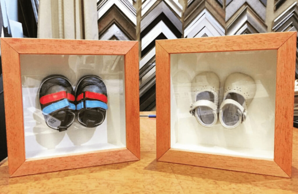 Dos marcos vitrina cuadrados de madera con profundidad que conservan y exponen los primeros zapatos de bebé: unas sandalias negras con velcro rojo y azul, y unos zapatos blancos de niña, recuerdos personales enmarcados por Contell Marcs Barcelona.