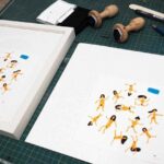Proceso de enmarcación en el taller Contell Marcs de una serigrafía con figuras humanas naranjas sobre papel blanco, junto a marco blanco listo para montar y sellos de caucho sobre tabla de corte verde.