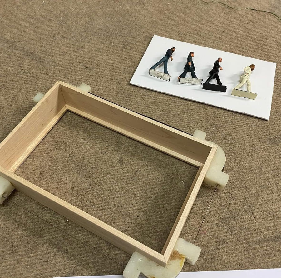 Caja de madera natural sin terminar con ensamblaje de esquinas junto a cuatro figuras en miniatura sobre papel blanco, proceso previo al montaje de vitrina diorama personalizada en Contell Marcs.