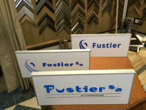 Tres rótulos corporativos de Fustier SA en diferentes tamaños sobre soporte blanco con logo azul, realizados por Contell Marcs con impresión digital y montaje adhesivado sobre forex y aluminio, expuestos en tienda de enmarcación con muestras de molduras al fondo.