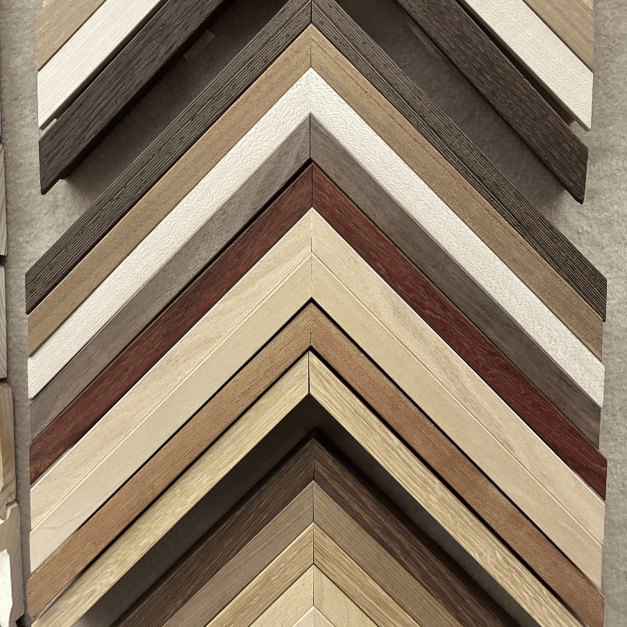 Panel de muestras de marcos de madera lisos apilados en abanico mostrando una amplia gama de tonos naturales: blanco, crema, beige, gris piedra, caoba rojizo y nogal oscuro, con veta visible, en el taller Contell Marcs de Barcelona.