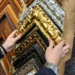 Manos de enmarcador del taller Contell Marcs sujetando una moldura de madera dorada tallada ante una amplia selección de marcos clásicos en acabados plateado, dorado, bronce oscuro, negro y madera natural, mostrando el asesoramiento personalizado del taller de Barcelona.