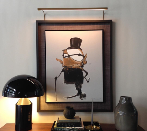 Ilustración de personaje con sombrero de copa, gafas y bastón enmarcada con marco de madera oscuro de moldura ancha, iluminada con foco de latón dorado, sobre consola de diseño con lámpara negra y objetos decorativos, trabajo realizado por Contell Marcs Barcelona.