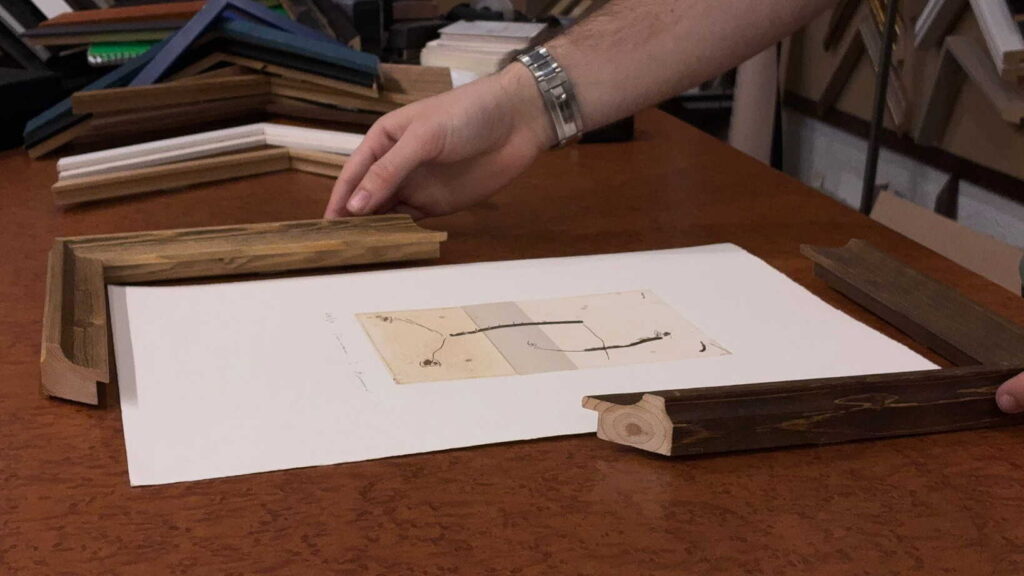 Manos de enmarcadora del taller Contell Marcs colocando una moldura de madera sobre una acuarela en papel blanco sobre mesa de trabajo, mostrando el proceso de asesoramiento y selección del marco más adecuado para la obra.