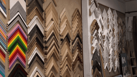 Vista interior del taller Contell Marcs de Barcelona con paredes repletas de centenares de molduras de marcos de madera apiladas en abanico, desde colores vivos hasta tonos naturales y acabados dorados y plateados, mostrando la amplia variedad disponible.